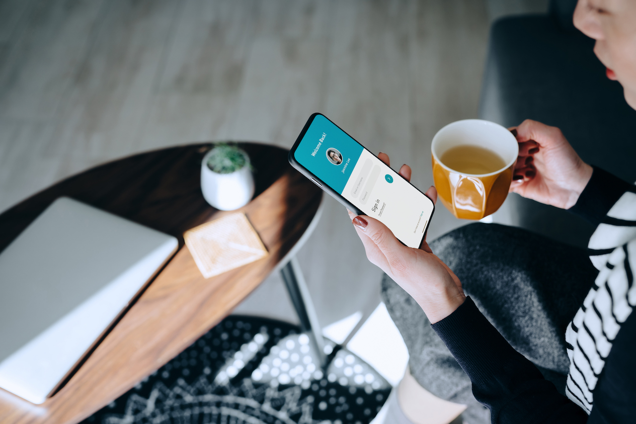 Young woman relaxing at home drinking a cup of tea, logging in to her smartphone device.