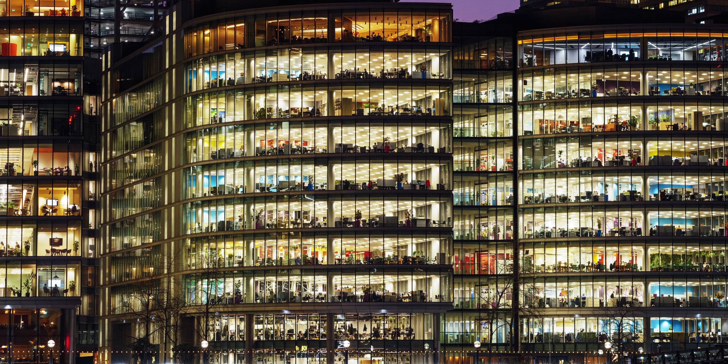 Illuminated office building at night with multiple floors of workers visible through floor-to-ceiling glass windows