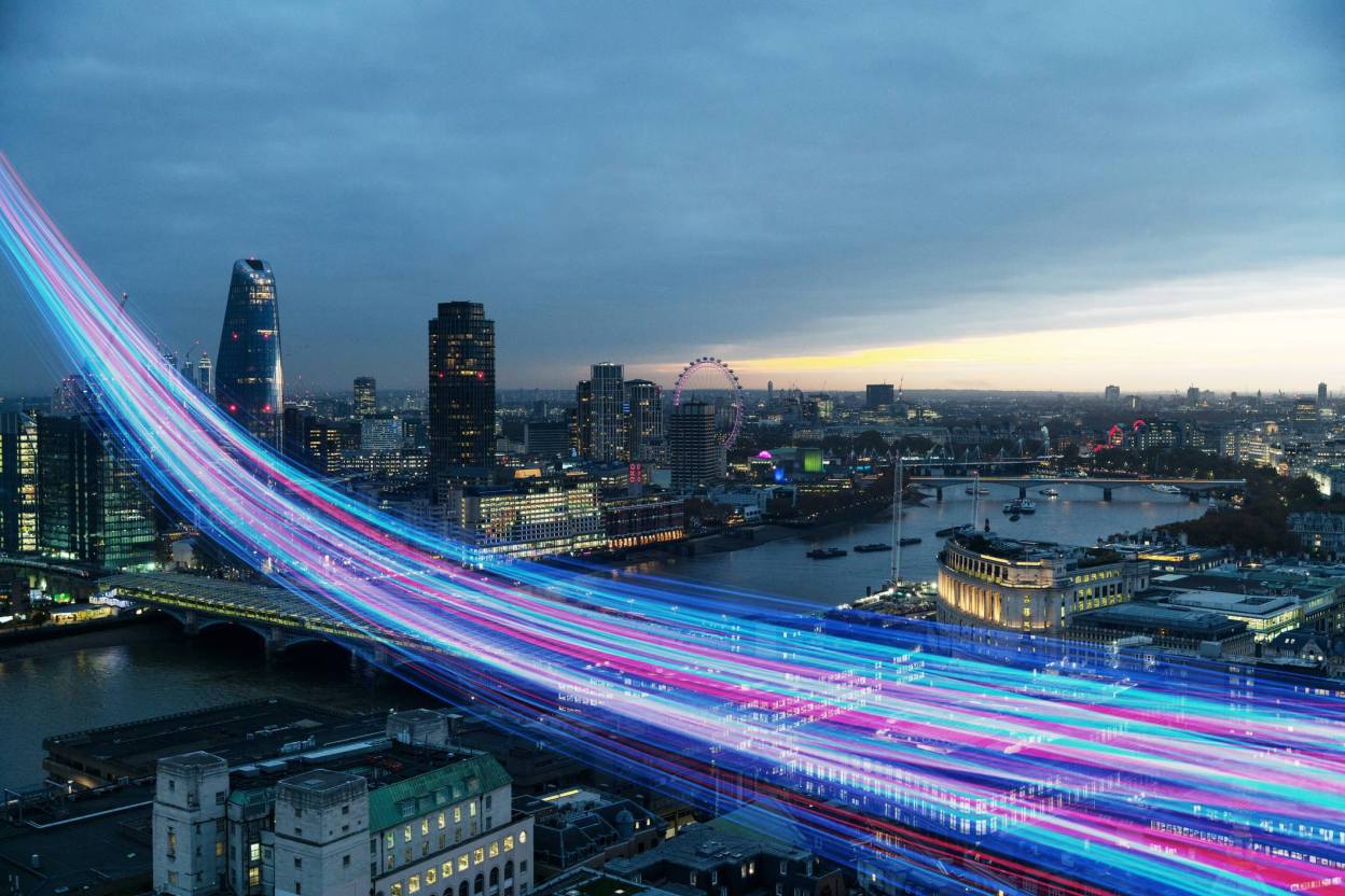 Aerial view of London skyline at dusk with glowing blue and pink digital data streams sweeping across the Thames, representing cloud automation velocity